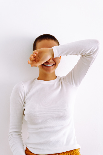 Beautiful slender laughing girl with shaved head short hairstyle, in white golf mock-ap, covers face with hand, hiding or embarrassed of looks