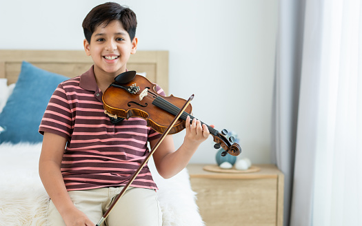 Indian handsome teenage boy, smiling, playing, practicing violin musical instrument with happiness in bedroom at cozy home in leisure time, smiling, having copy space. Education, Lifestyle, Creativity Concept
