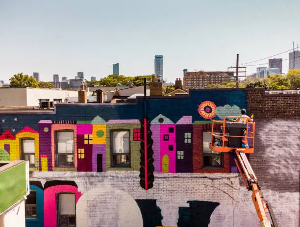 Aerial view of female artist painting large wall mural from the crane. She is dressed in casual work clothes. Exterior of public  downtown of large North American City. Captured from the air via drone.