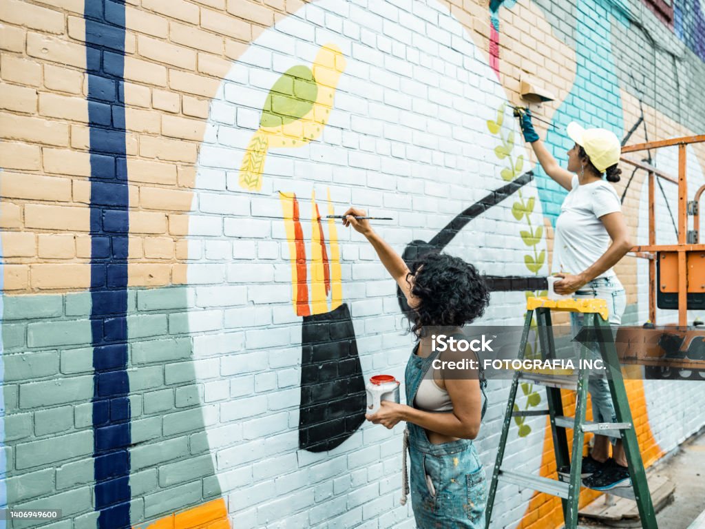Two Female artists painting large wall mural Two Female artists painting large wall mural. They are dressed in casual work clothes. Exterior of public  downtown of large North American City. Captured from the air via drone. Art Stock Photo