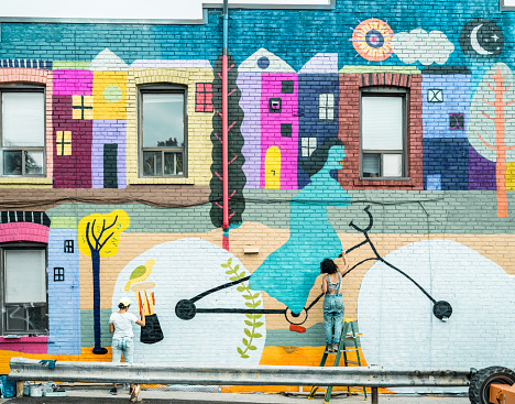 Female artist painting large wall mural. She is dressed in casual work clothes. Exterior of public  downtown of large North American City. Captured from the air via drone.