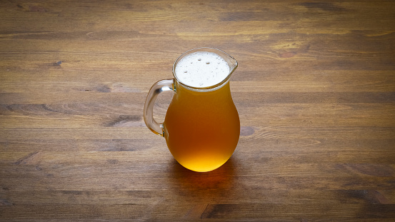 A golden pint of beer with foam at the top of the glass.  The glass of beer is placed on a hardwood surface with liquid drops of beer surrounding the glass on the table.