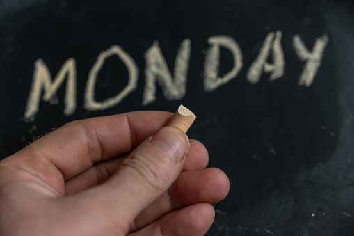 Adult male holding a piece of yellow chalk in his hand. Handwritten word MONDAY on black chalkboard. Days of the week. Calendar