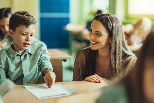 Woman in glasses and positive children examining globe while standing around table in geography classroom during lesson at school