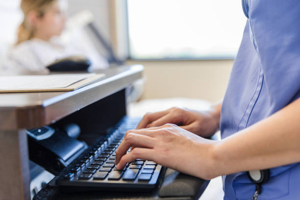 Focus on healthcare worker's hands typing on computer keyboard The focus of the photo is on the healthcare worker's hands as she types on the computer keyboard.  An unrecognizable female patient waits quietly in the background. electronic medical record stock pictures, royalty-free photos & images