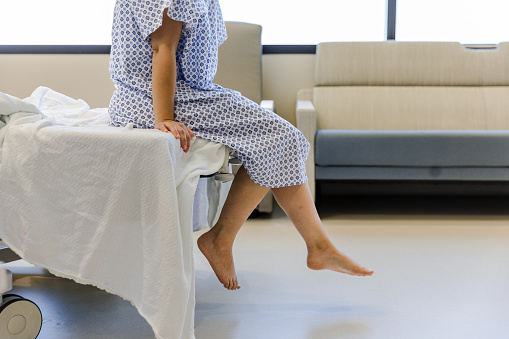 An unrecognizable female patient wears a hospital gown and sits on the edge of the hospital bed.  She dangles her feet while waiting for her test results.