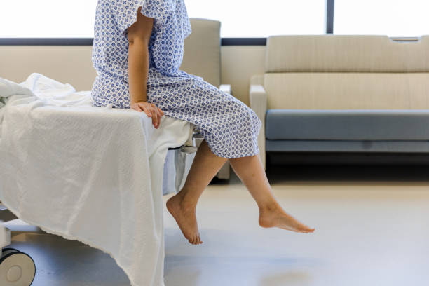 Unrecognizable patient in hospital gown waits for test results An unrecognizable female patient wears a hospital gown and sits on the edge of the hospital bed.  She dangles her feet while waiting for her test results. gynaecologist stock pictures, royalty-free photos & images