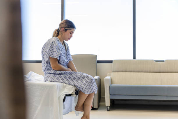 ansiosa, triste, joven mujer vestida con bata de hospital mira hacia abajo - examination gown fotografías e imágenes de stock