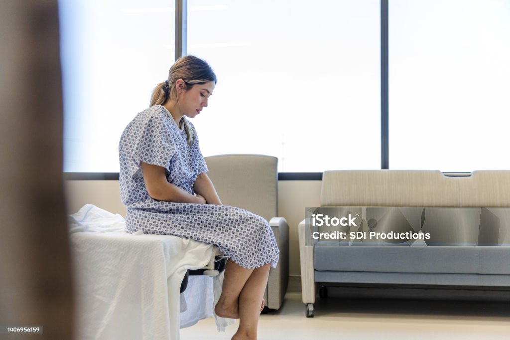 Anxious, sad, young woman wearing hospital gown looks down The anxious, sad, young female patient wears her gown as she waits in the hospital room. Hospital Stock Photo