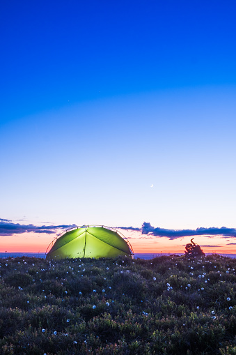 Dome tent illuminated at sunset beside bikepacking bike on mountain ridge overlooking valley below crescent moon.