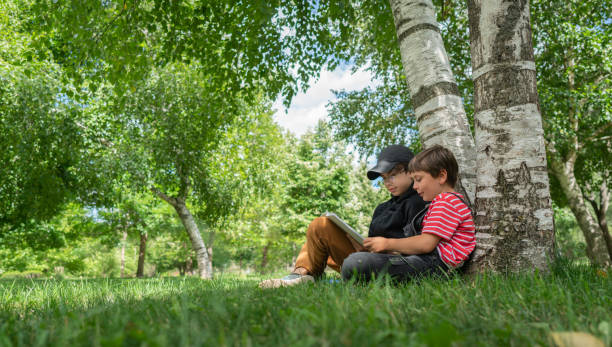 Two school kid boys sitting under a tree and read books on a sunny summer day