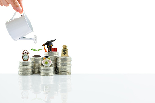 Saving money for college tuition fee concept : Hand pours water from watering can, clock, sprout / small tree, US dollar bag, book and black cap on steps of coins, isolated on white background.