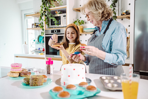 Cute little girl helping her grandmother in the kitchen. They are having fun while decorating cake