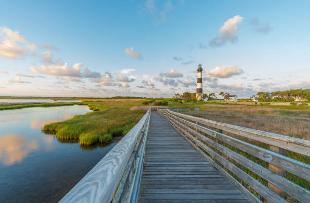 Lighthouse with Boardwalk Through Marsh at Bodie Island, NC. The lighthouse at Bodie Island has been in operation as a navigational aid since October 1, 1872. Its powerful light beams warn sailors traversing the treacherous area known as “Graveyard of the Atlantic.  It is now operated by the National Park Service. cape hatteras stock pictures, royalty-free photos & images