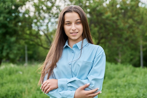 Outdoor portrait of a beautiful teenage girl 15 years old. Smiling female teenager looking at the camera in the park, against the backdrop of green grass. Adolescence, high school, age concept