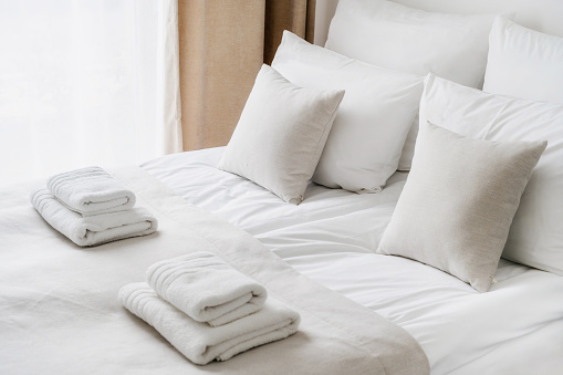 Cropped view of white bedclothes and towels on bed in comfortable hotel room, hypoallergenic pillows, personal comfort idea, bedding concept, bathroom details