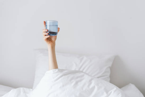 Woman holding glass of water after awakeness Person's hand holding glass of water when lying in bed, morning rituals, copy space, morning in contemporary hotel room, wellness and relax, purified water after party stock pictures, royalty-free photos & images