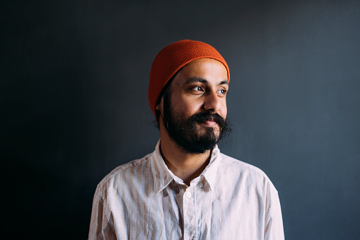 A portrait of an Indian influencer fashionably dressed smiling while looking away with an orange cap on his head.