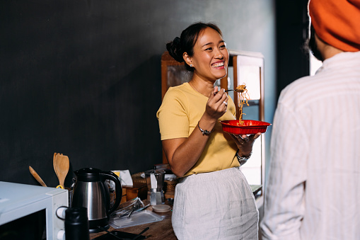 A happy Asian female eating pasta from a red takeaway food container while having a conversation with an anonymous man.