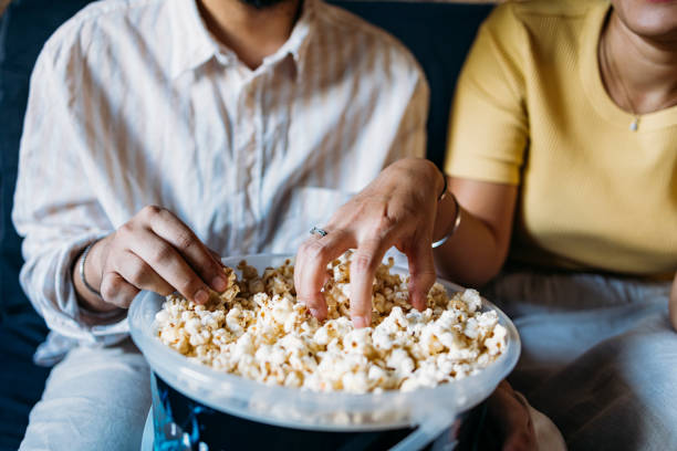 An Unrecognizable Mixed-race Couple Enjoying Eating Popcorn While Relaxing At Home An anonymous Asian woman and her Indian boyfriend eating snacks while spending their leisure time together. Plain Popcorn stock pictures, royalty-free photos & images