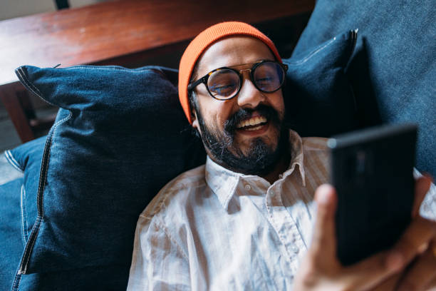 un hombre guapo y feliz con gafas y una gorra naranja en la cabeza sosteniendo su teléfono móvil mientras está acostado en el sofá - telecommunications equipment discussion telephone horizontal fotografías e imágenes de stock