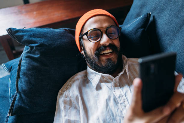 szczęśliwy przystojny mężczyzna w okularach i pomarańczowej czapce na głowie trzymający telefon komórkowy leżąc na sofie - wireless technology working at home one person reading zdjęcia i obrazy z banku zdjęć