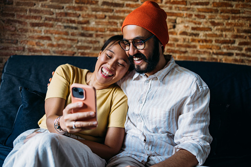A beautiful smiling Asian woman and her Indian boyfriend reading something on social media using an app on the smartphone while sitting on the sofa in the living room.