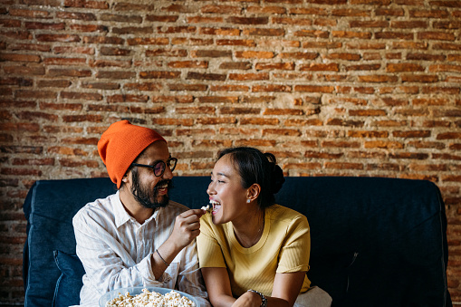 A smiling Asian woman and her Indian boyfriend spending their leisure time together while sitting on the sofa in the living room eating snacks.