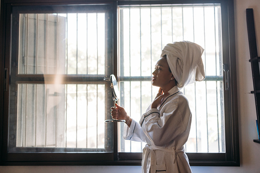 A side view of a confident Asian female in bathrobe standing near the window and thinking about the most suitable cosmetic product to use after the shower.