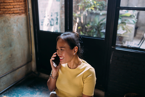 A smiling Asian female freelancer having a conversation with somebody using her smartphone.