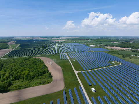 A high aerial view above a rural area that is filled with solar panels, a vast solar farm filling the landscape on a partly cloudy summer's day.