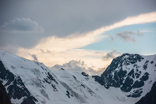 Dramatic aerial landscape with large snowy mountain range under cloudy sky in golden sunset sunlight. Awesome alpine scenery with snow mountains under gold clouds in blue sky at changeable weather.