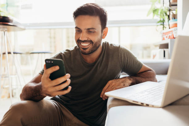 hombre alegre usando teléfono inteligente y computadora portátil en casa - teléfono móvil fotografías e imágenes de stock