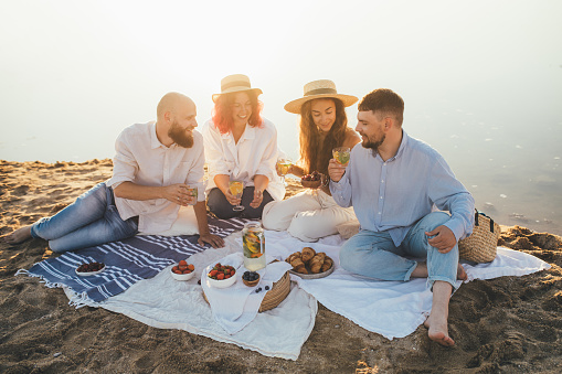Group of happy friends having picnic, drinking lemonade and eating fruits, sitting near sea at sunset. Summer vacation.