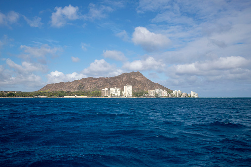 Diamond Head is an iconic volcanic cone in Waikiki, Hawaii.  This mountain is often the symbol of Honolulu.  This view is taken from the ocean.  The water is deep blue.  The coast line is known as the gold coast of Waikiki.  Here you can see beach goers and surfers in the distance.  A few scattered, fluffy clouds float by.