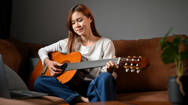 женщина расслабляется, сидя в гостиной, играя на своей акустической гитаре. - 20s acoustic guitar adult attractive female стоковые фото и изображения