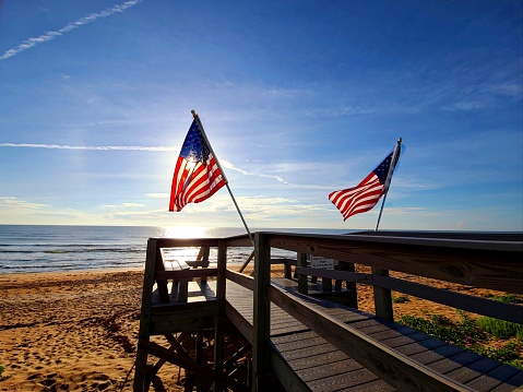 Two American flags resting against a wooden walkway over the beach with the ocean behind them