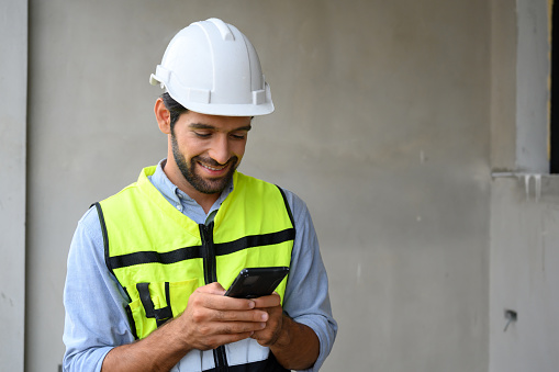 portrait of young engineer in vest with white helmet standing on construction site, smiling and holding smartphone for worker, internet, social media
