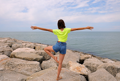 young slender girl on the rocks of the dike near the sea while performing gymnastic exercises to keep balance with one leg