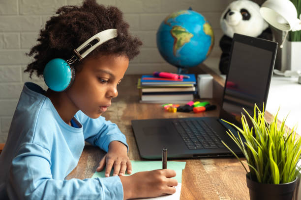 African-American girl doing homework,making notes,using headphones and laptop at home.Back to school concept.School distance education at home,home schooling,diverse people