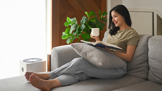 Pleasant young asian woman resting on comfortable sofa and reading book, spending leisure weekend at home.