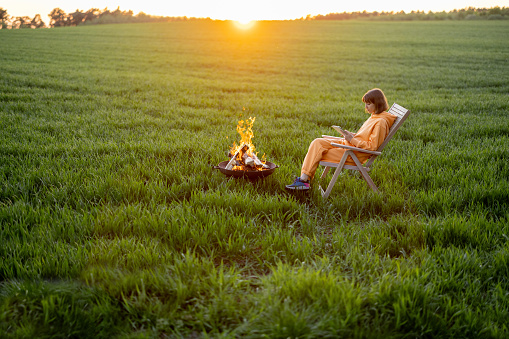 Young person sitting on chair near the fireplace and using digital device while relaxing on green field during the sunset. Concept of recreation on nature and freelance