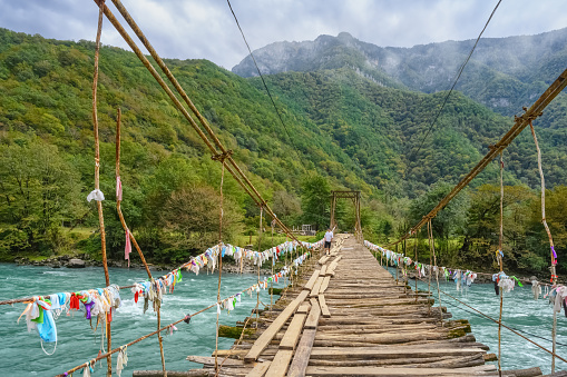 A dilapidated suspension bridge across the mountain river Bzyb in Abkhazia. Old floor in the form of separate boards. Tourists tie colorful knots to metal handrails for good luck. Tour to Lake Ritsa.