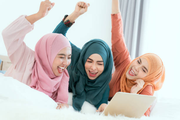 sekelompok wanita muslim berbaring di tempat tidur mengobrol dengan gembira di laptop mereka. - cantik hijab potret stok, foto, & gambar bebas royalti