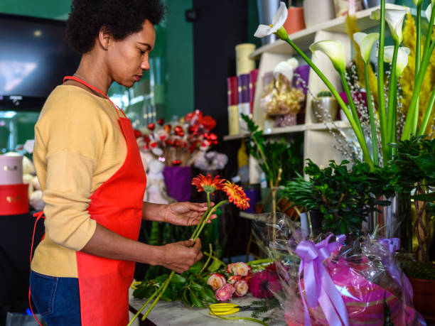afroamerikanische frau in einer schürze, die mit blumen arbeitet und einen blumenstrauß arrangiert - rose flower shop variation color image stock-fotos und bilder