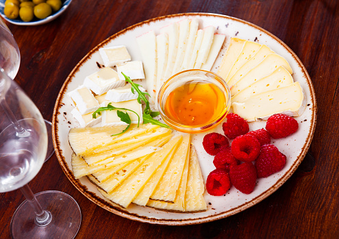 Different types of cheese sliced and served on plate with little bowl of honey and marinated olives.