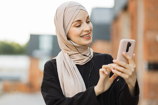 Portrait of young modern arabian woman holding mobile phone and listening the music to headphone. Muslim woman with smartphone and headphones walking on a city street. Pretty girl in hijab on sidewalk