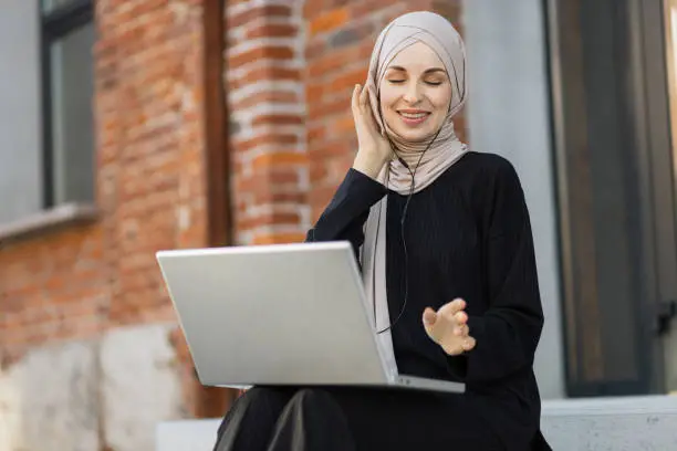 Music streaming platform concept. Joyful muslim girl in hijab wearing wireless headphones, listening favorite songs online working on laptop, relaxing during coffee break, on city background.