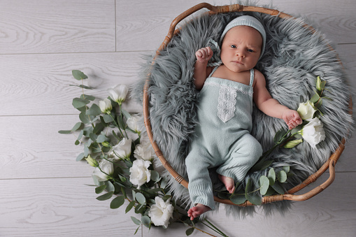 Adorable newborn baby lying in basket with faux fur and flowers on floor, top view