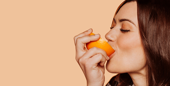 Close-up portrait of young cheerful woman, eating orange with eyes closed. Face shot at studio over beige background.